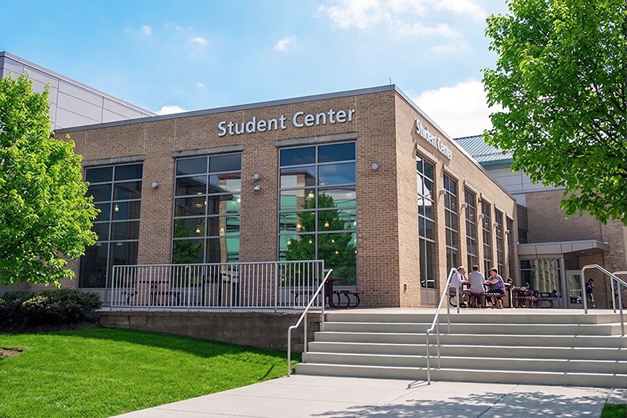 A bright, brick student center building features large windows and a concrete staircase leading to an outdoor patio with metal railings. Lush green trees and a grassy lawn surround the modern structure under a clear blue sky.