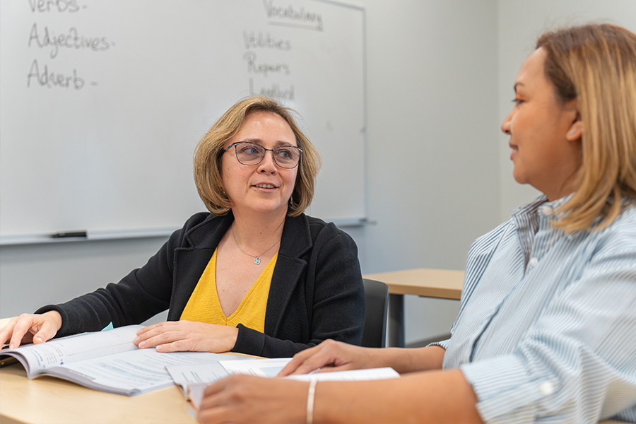 A faculty member wearing glasses and a black cardigan over a yellow top sits at a desk, smiling and talking with an adult student.