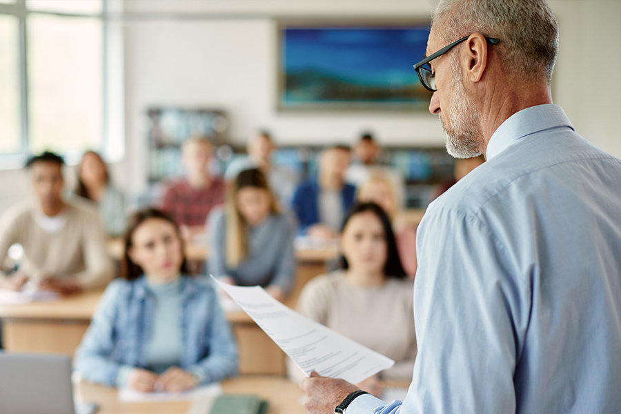A professor holding papers addresses a classroom full of attentive students. A professor holding papers addresses a classroom full of attentive students.