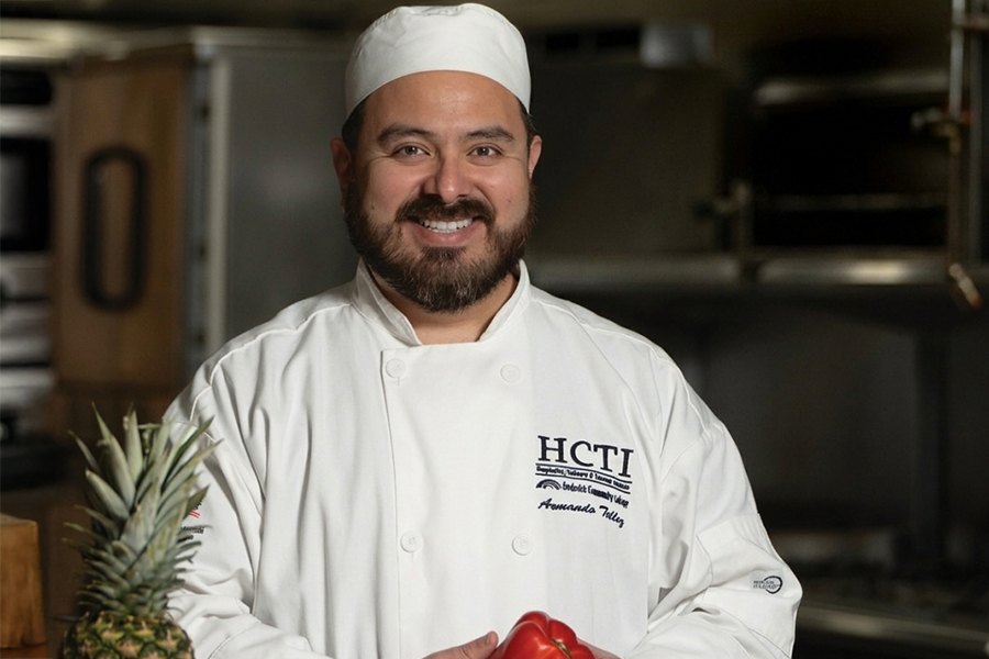 A smiling FCC HCTI chef in a white chef's coat holding fresh produce in a commercial kitchen.