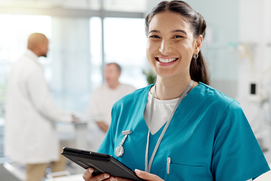 A smiling female healthcare professional in teal scrubs and a badge reel holding a digital tablet. In the blurred background, other medical staff in white lab coats are working in a clinical setting.