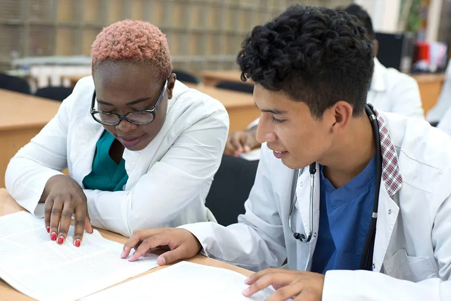 Two students in white lab coats and stethoscopes lean over an open textbook together, reviewing material in a classroom setting.