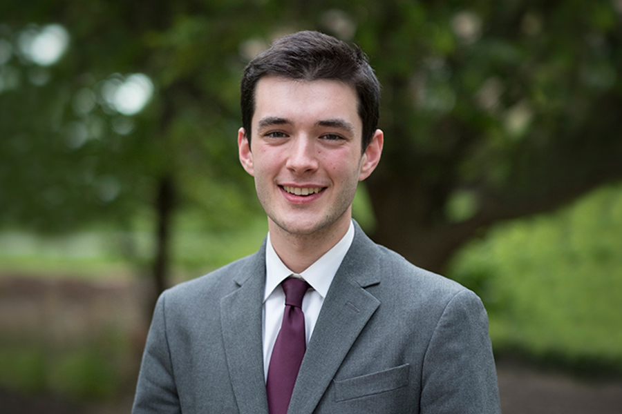 A young man in a gray suit and burgundy tie smiles outdoors with green trees in the background.