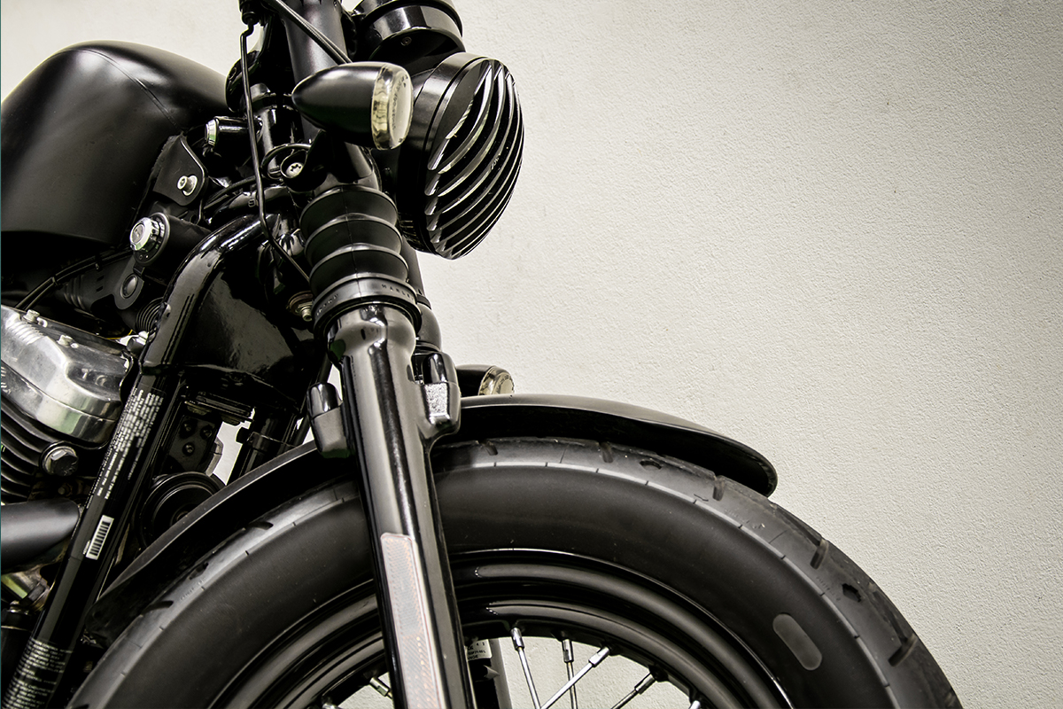 Close-up of the front wheel, fork, and headlight of a black motorcycle against a light gray background.