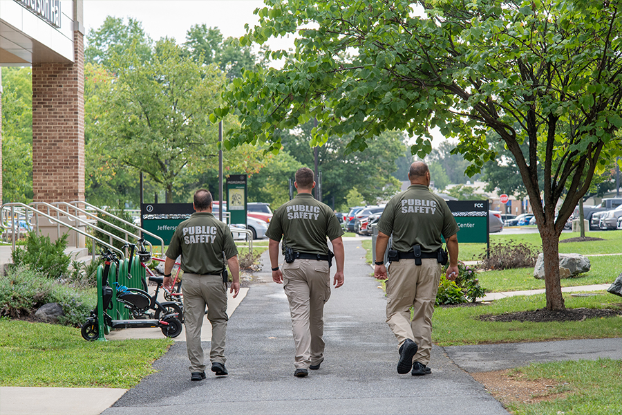 Three Frederick Community College Public Safety officers in olive green uniforms and khaki pants walk together down a paved campus pathway toward the campus center.