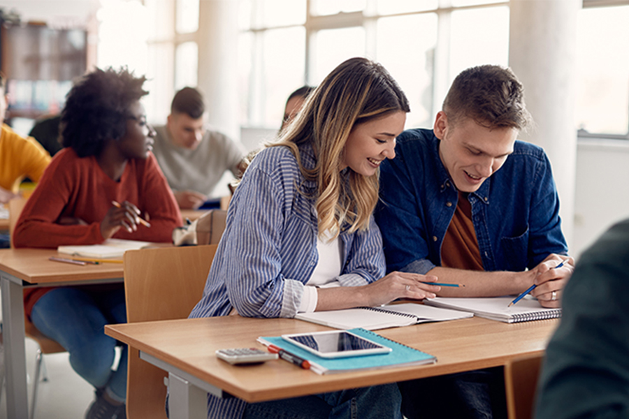 Two college students, a woman in a blue striped shirt and a man in a denim shirt, sit together at a desk in a classroom, smiling while looking at a notebook. They appear to be collaborating on an assignment. In the background, other students are visible at their desks, including a woman in a burnt-orange sweater.