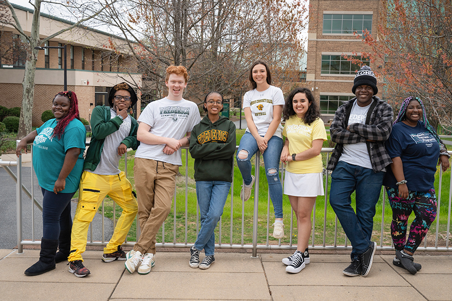 Eight diverse students leaning against a metal railing on a college campus, smiling and posing in a relaxed, friendly manner. They are wearing a mix of casual clothing and college-branded apparel, with academic buildings and trees in the background.