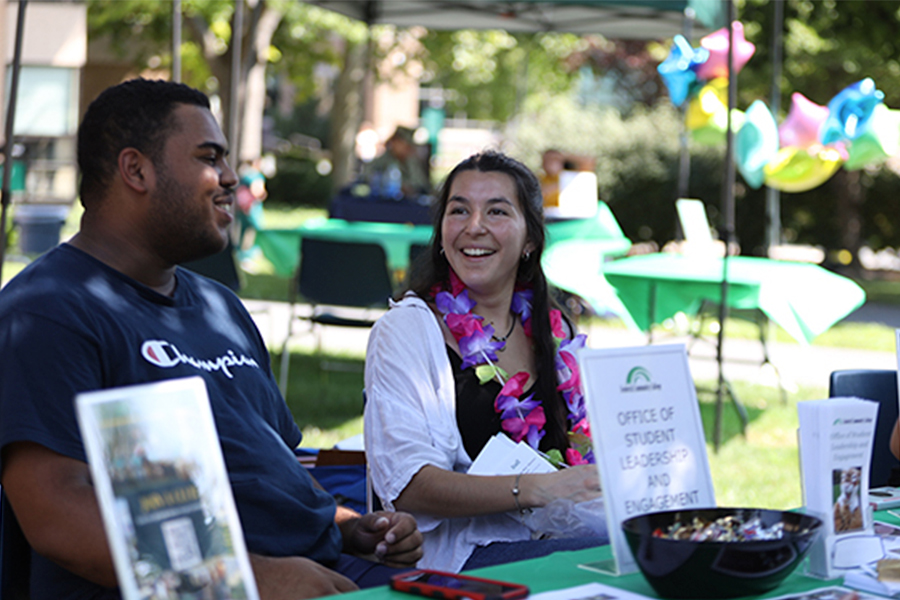 Two students smiling and talking at an outdoor "Office of Student Leadership and Engagement" information table.