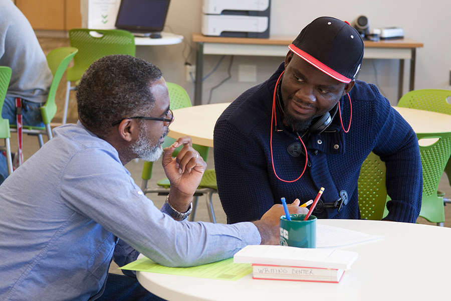 Two men engage in a focused tutoring session at a round table in a modern learning center. The man on the left, wearing glasses and a blue shirt, explains a point while the man on the right, wearing a black cap and headphones, listens intently.
