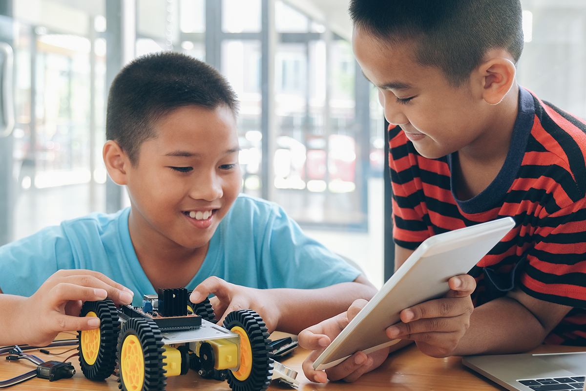 Two boys collaborate on building a wheeled robotics vehicle, with one adjusting the model and the other holding a tablet. They smile as they work together at a table.