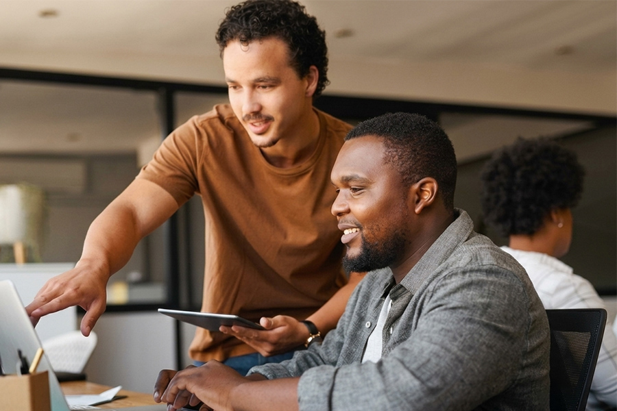 Two men collaborate at a desk, with one standing and pointing at a laptop screen while the other looks on and smiles.