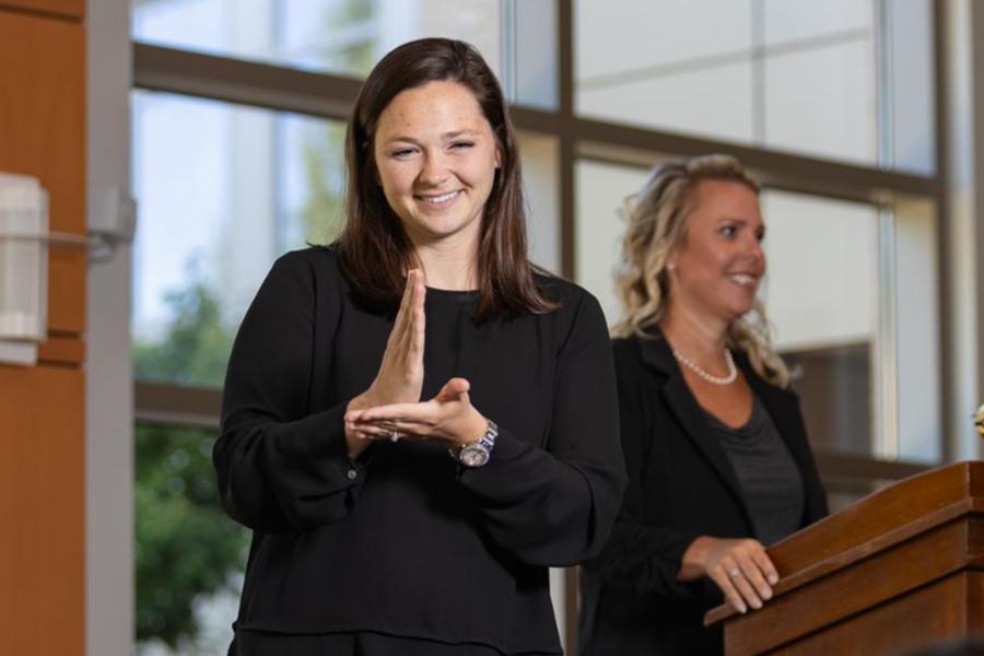 A sign language interpreter in a black blouse provides interpretation during a presentation or event, with a speaker standing at a wooden podium in the background.