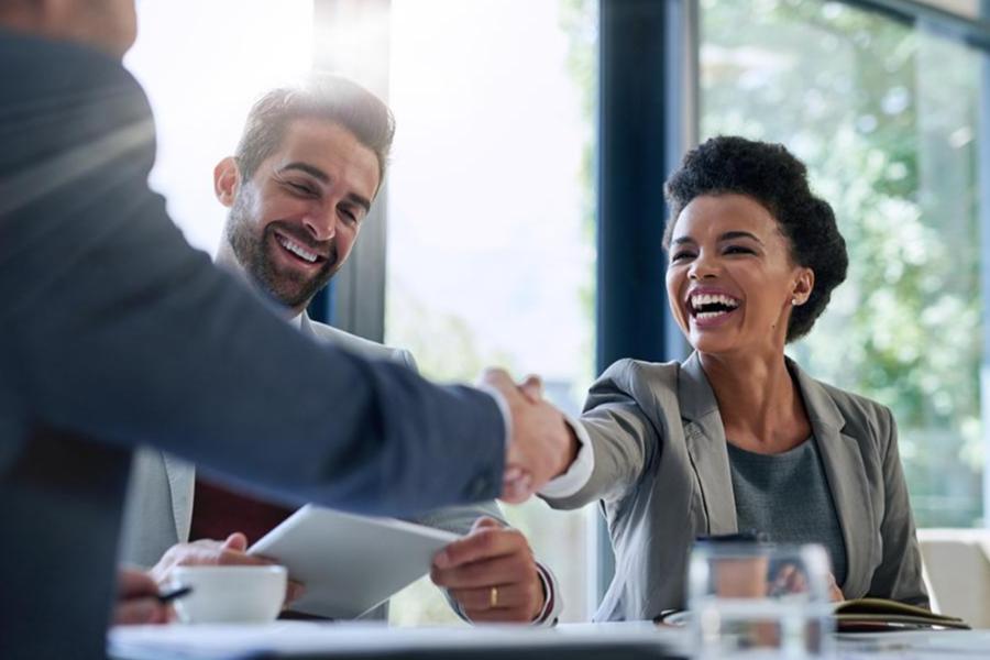 A smiling woman in a grey blazer shakes hands with a colleague across a table during a professional meeting.