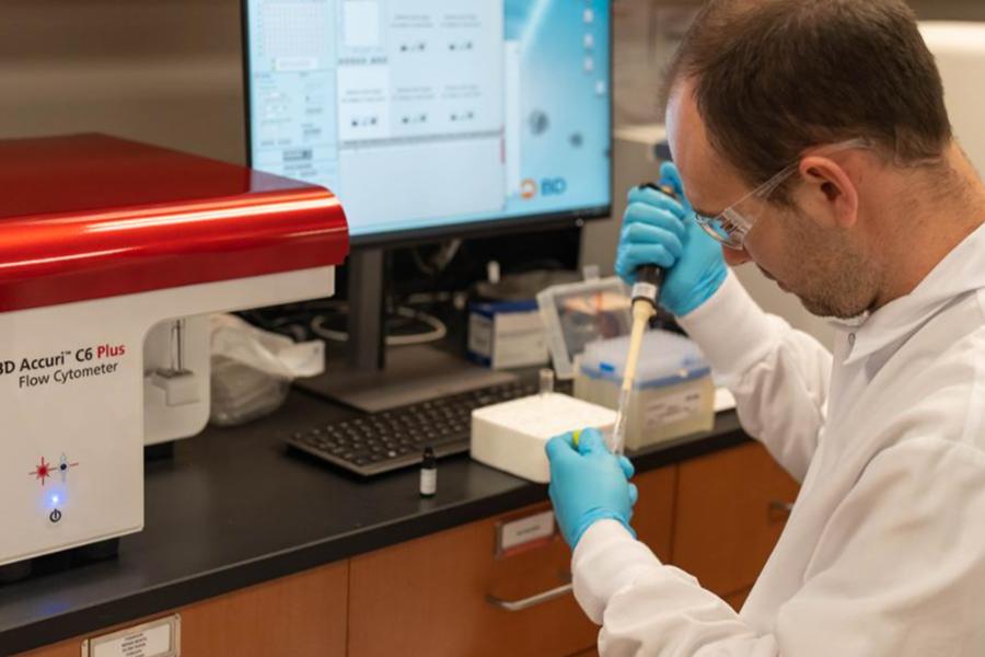 A scientist in a lab coat and gloves uses a pipette beside a BD Accuri C6 Plus Flow Cytometer. 