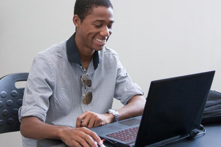 A young man with a smile sits at a desk and uses a laptop with an external mouse.