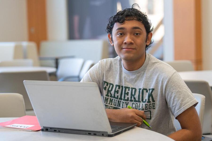 A young man wearing a Frederick Community College t-shirt sits at a table with an open laptop and a pen.