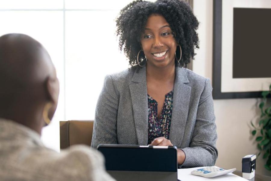 Gemini said A smiling woman with curly black hair and hoop earrings is sitting at a desk, engaging in a conversation with someone seated across from her (seen from the back). She is wearing a grey blazer over a patterned blouse. On the desk in front of her are a tablet in a stand, a calculator, and some paperwork, suggesting a professional consultation or meeting in an office setting.