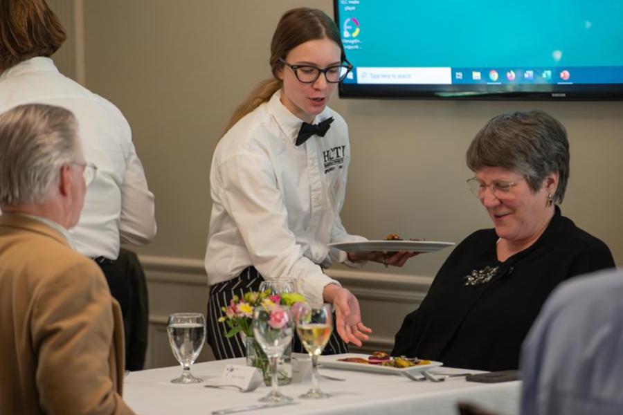 A culinary student in a white server's uniform and black bowtie presents a plated meal to a guest in a formal dining setting.