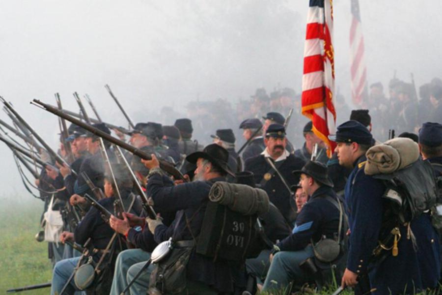 A group of historical reenactors dressed in American Civil War Union uniforms sit and stand in a foggy field, holding period-accurate rifles and a large American flag.