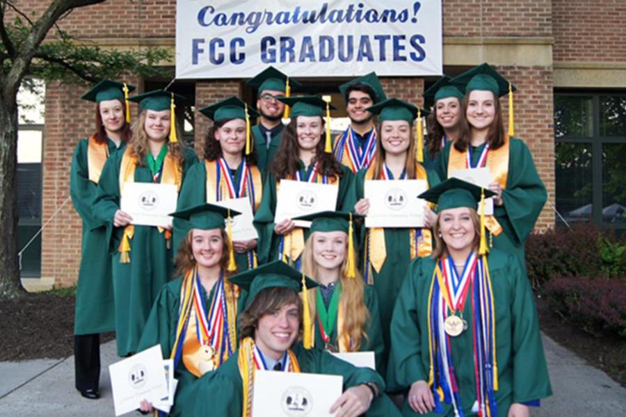 A group of graduates in green caps and gowns hold their diplomas and wear honor cords in front of a brick building with a "Congratulations! FCC Graduates" banner.