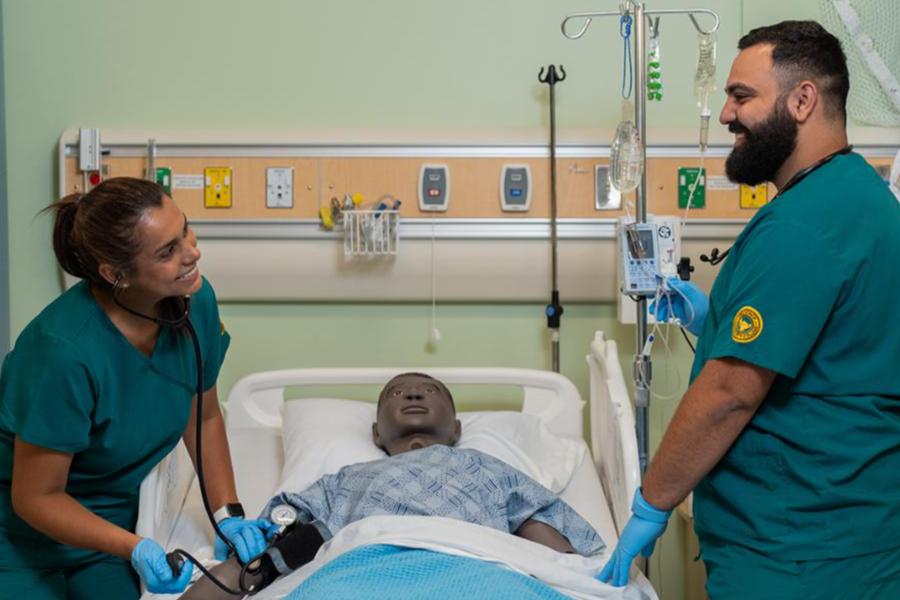 Two nursing students in green scrubs practice clinical skills on a medical mannequin in a simulated hospital room equipped with IV stands and monitoring equipment.