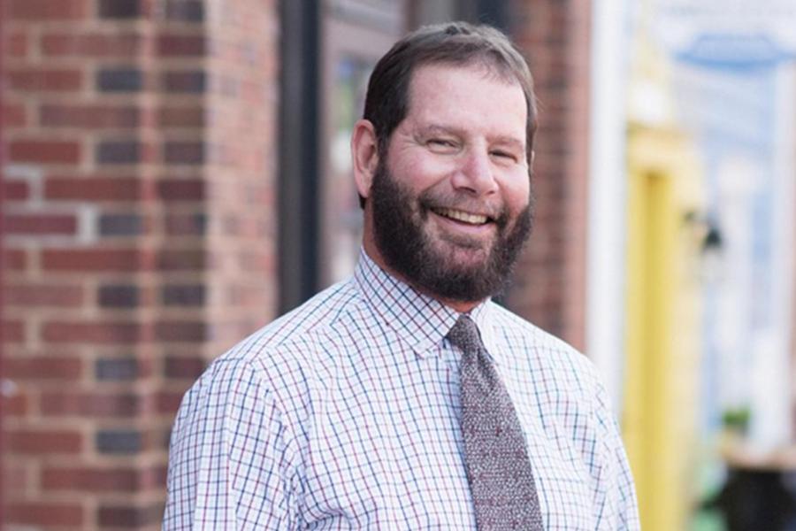 A smiling middle-aged man with a beard wearing a button-down shirt and tie standing outdoors.