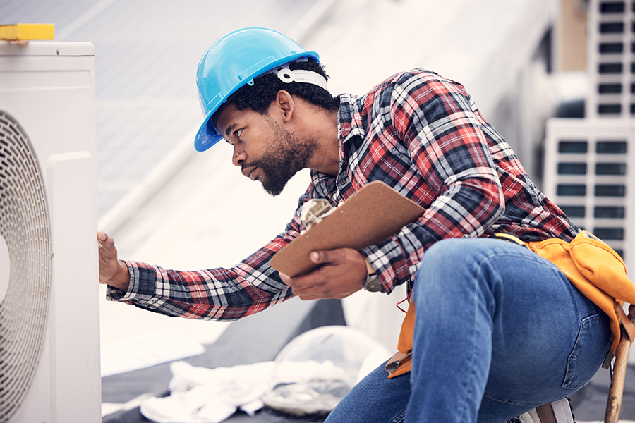 An HVAC technician wearing a blue hard hat and holding a clipboard inspects an outdoor rooftop air conditioning unit.