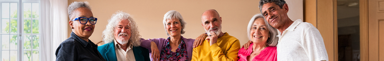 A diverse group of six older adults smile and pose together with their arms around each other in a bright, warmly lit indoor space.