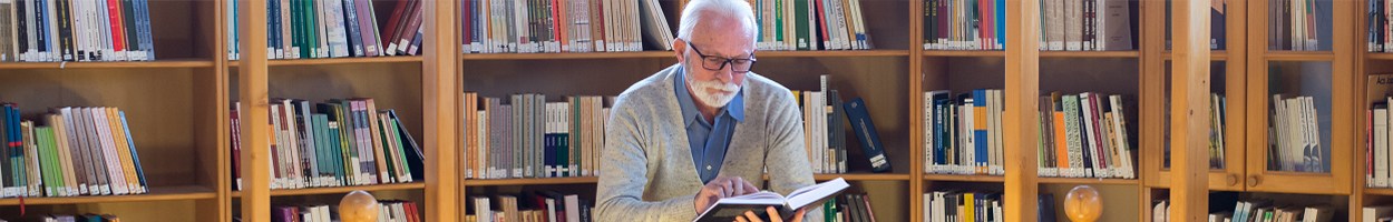 An older man with white hair and a beard reads an open book while seated in front of tall wooden bookshelves filled with books in a library.