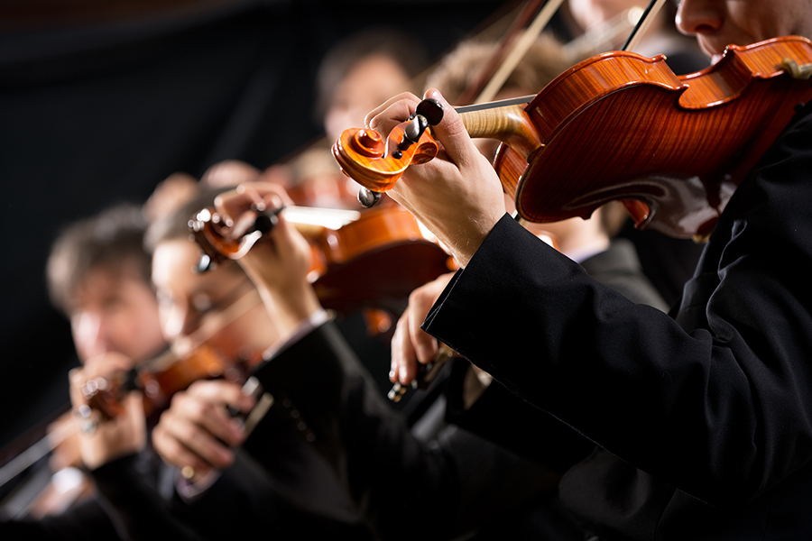 A close-up, side-angle view shows several violinists in black formal wear playing their instruments in an orchestra.