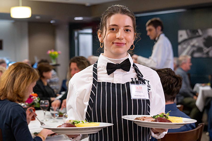 Culinary student in a bow tie and striped apron holds two plated dinners in a dining room